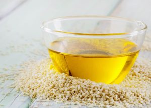 A Glass Bowl Filled With Yellow Oil, Surrounded By Sesame Seeds On A Light Wood Surface.