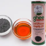 A Bottle Of Ghee Store Cold Pressed Gingelly Oil Is Placed Beside A Bowl Of Black Sesame Seeds And A Bowl Of The Oil.