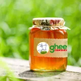 A Jar Of Raw Wild Honey With A Decorative Lid, Labelled &Quot;Ghee Store&Quot; With A Cow Graphic, Placed Outdoors On A Wooden Surface With Greenery In The Background.