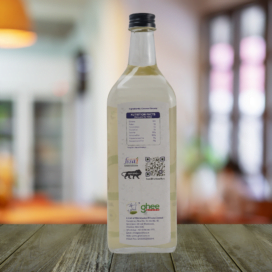 Back View Of A Clear Bottle With Nutritional Facts Label And Contact Information On A Wood Surface, Blurred Indoor Setting In The Background.